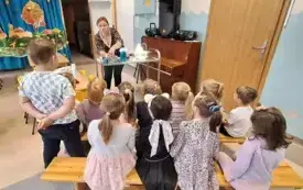 dzieci obserwują eksperymenty kt&oacute;re wykonuje pani nauczycielka stojąca za metalowym stolikiem na kt&oacute;rym stoją r&oacute;żne pojemniki 