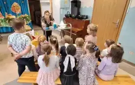 dzieci obserwują eksperymenty kt&oacute;re wykonuje pani nauczycielka stojąca za metalowym stolikiem na kt&oacute;rym stoją r&oacute;żne pojemniki 