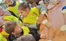 dzieci w kamizelkach odblaskowych siedzą przy stole na kt&oacute;rym stoją plastikowe kubeczki 
