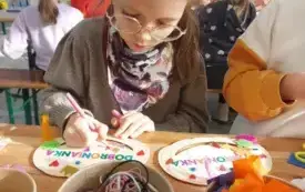 dziewczynka w okularach dekoruje szablon dyni za pomocą flamastra przed nią stoją w pudełkach kolorowe sznurki i pi&oacute;rka