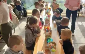 dzieci siedzą wzdłuż długiego stołu przed nimi na stole stoją r&oacute;żne przybory plastyczne