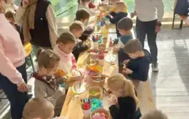 dzieci siedzą przy długim stole na kt&oacute;rym leżą r&oacute;żne ozdoby plastyczne i szablony z drewna dynia z napisem dobronianka
