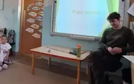pan leśnik siedzi na krześle obok tablica multimedialna na kt&oacute;rej wyświetlony jest napis praca leśnika