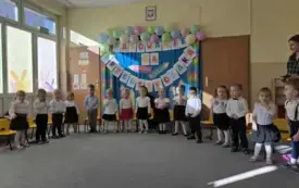 dzieci w odświętnych ubraniach siedzą na krzesełkach w p&oacute;łkolu za nimi dekoracja z napisem Pasowanie na przedszkolaka szablony kredek i balony po prawej stronie stoi pani