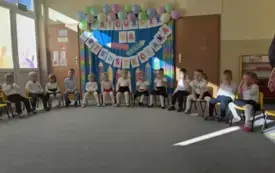 dzieci w odświętnych ubraniach siedzą na krzesełkach w p&oacute;łkolu za nimi dekoracja z napisem Pasowanie na przedszkolaka szablony kredek i balony