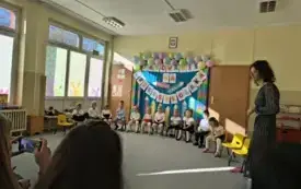 dzieci w odświętnych ubraniach siedzą na krzesełkach w p&oacute;łkolu za nimi dekoracja z napisem Pasowanie na przedszkolaka szablony kredek i balony obok stoi pani nauczycielka