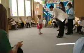 dzieci w odświętnych ubraniach siedzą na krzesełkach w p&oacute;łkolu za nimi dekoracja z napisem Pasowanie na przedszkolaka szablony kredek i balony na środku stoją trzy panie jedna w białej marynarce trzyma dużą kredkę nad odświętnie ubraną dziewczynką