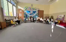 dzieci w odświętnych ubraniach siedzą na krzesełkach w p&oacute;łkolu za nimi dekoracja z napisem Pasowanie na przedszkolaka szablony kredek i balony