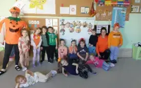 dzieci pozują do zdjęcia z paniami z kt&oacute;rych jedna jest w pomarańczowej bluzie a druga w przebraniu dyni dzieci mają w stroju akcenty dyniowe lub hallowenowe