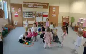 dzieci wraz z panią stoją na dywanie na kt&oacute;rym leżą dwa okręgi a w nich kolorowe piłeczki dwoje dzieci kuca obok okręg&oacute;w