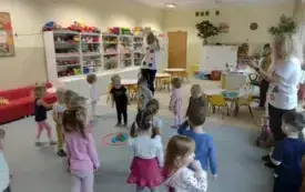 dzieci wraz z trzema paniami stoją na dywanie na kt&oacute;rym leży okręg a w nim kolorowe piłeczki