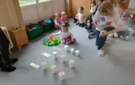 dzieci siedzą skupione na dywanie dziewczynka sięga po piłeczkę do okręgu w kt&oacute;rym jest dużo kolorowych piłeczek obok klęczy pani przed nimi leżą karteczki z kolorowymi k&oacute;łkami i stoją plastikowe kubki