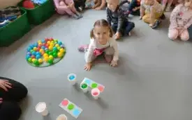 dzieci siedzą skupione na dywanie dziewczynka siedzi przed obrazkami z kolorowymi k&oacute;łkami i piłeczkami w plastikowych kubeczkach obok w kręgu leży wiele kolorowych piłeczek