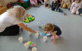 dzieci siedzą skupione na dywanie dziewczynka wraz z panią kucają przed obrazkami z kolorowymi k&oacute;łkami dziewczynka wkłada piłeczkę do plastikowego kubka obok w kręgu leży wiele kolorowych piłeczek