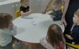 dzieci siedzą przy okrągłym stoliku na kt&oacute;rym leży sylweta twarzy nad nimi pochyla się pani
