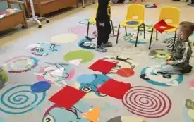 dzieci stoją i siedzą na dywanie i obok niego widać krzesełka ustawione w szeregi po trzy  sztuki na dywanie leżą kolorowe figury geometryczne