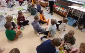 dzieci siedzą w małych grupach na kolorowym dywanie pomiędzy nimi stoją kolorowe kubeczki