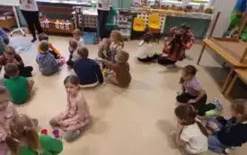 dzieci siedzą w małych grupach na kolorowym dywanie pomiędzy nimi stoją kolorowe kubeczki