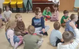 dzieci siedzą w małych grupach na kolorowym dywanie pomiędzy nimi stoją kolorowe kubeczki