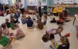 dzieci siedzą w małych grupach na kolorowym dywanie pomiędzy nimi stoją kolorowe kubeczki