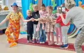 sz&oacute;stka dzieci stoi w rzędzie  i trzymają na rękach węża obok kuca pani w kolorowym ubraniu i niebieskim kapeluszu 