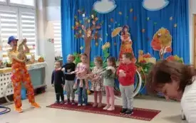 sz&oacute;stka dzieci stoi w rzędzie obok stoi pani w kolorowym ubraniu i niebieskim kapeluszu i trzyma węża