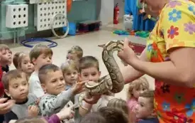 pani w kolorowym ubraniu i niebieskim kapeluszu trzyma węża na rękach i pozwala dotknąć go dzieciom