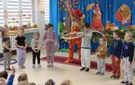 pani w kolorowym ubraniu i peruce wraz z dziećmi stoją w p&oacute;łkolu pani m&oacute;wi coś do mikrofonu dwoje dzieci trzyma obręcze