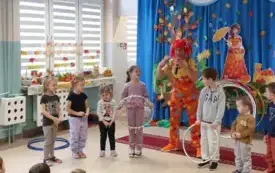 pani w kolorowym ubraniu i peruce wraz z dziećmi stoją w p&oacute;łkolu pani m&oacute;wi coś do mikrofonu dwoje dzieci trzyma obręcze
