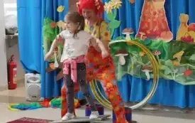 pani w kolorowym ubraniu i peruce asekuruje dziewczynkę stojącą na na r&oacute;wnoważni 