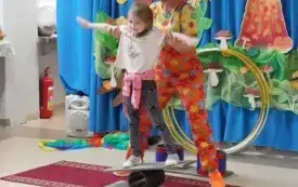 pani w kolorowym ubraniu i peruce asekuruje dziewczynkę stojącą na na r&oacute;wnoważni 