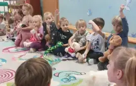 dzieci siedzą w szeregu niekt&oacute;re podnoszą rękę do g&oacute;ry dzieci trzymaja pluszowe zabawki