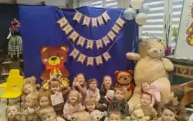 dzieci z pluszakami w rękach siedzą na dywanie za nimi dekoracja dzień pluszowego misia