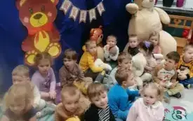 dzieci z pluszakami w rękach siedzą na dywanie za nimi dekoracja dzień pluszowego misia