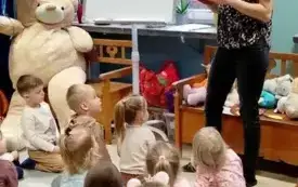 pani stoi przed dziećmi kt&oacute;re siedzą na dywanie obok pani na tablicy wisi duża sylwetka misia kt&oacute;rą pani układa