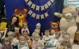 dzieci wraz z panią siedzą na dywanie i pozuja do zdjęcia na tle dekoracji z misiami i napisem dzień pluszowego misia pani ma założoną opaskę z uszami misia