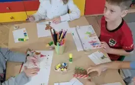 dzieci siedzą przy stole na kt&oacute;rym leżą rysunki i stoją kredki w pojemniku