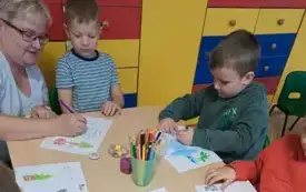 dw&oacute;ch chłopc&oacute;w pochylonych nad rysunkami obok kuca pani i pomaga jednemu z chłopc&oacute;w na stole stoją kredki w pojemniku