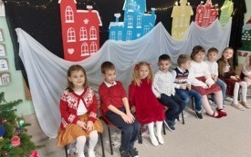 dzieci siedzą obok siebie na krzesełkach za nimi dekoracja zimowe kolorowe domki dzieci w świątecznych ubraniach