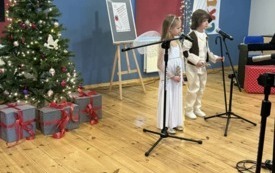dziewczynka w stroju aniołka chłopiec w stroju pastuszka stoją przed mikrofonem obok stoi choinka widać rozstawione statywy do mikrofon&oacute;w