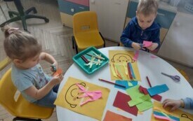 dwoje dzieci siedzi przy stoliku przed nimi leżą kolorowe kartki i ż&oacute;łte kartki z narysowaną buzią dzieci przyklejają kolorowe paski do kartek z buziami w miejscu włos&oacute;w