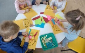 dzieci siedzą przy stoliku przed nimi leżą kolorowe kartki i ż&oacute;łte kartki z narysowaną buzią