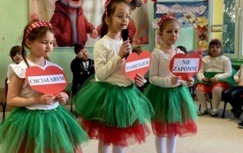 dzieci w odświętnych ubraniach siedzą na krzesełkach w p&oacute;łkolu za nimi dekoracja na dzień babci i dziadka trzy dziewczynki stoją na środku trzymają duże czerwone serca z napisami 