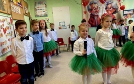dzieci w odświętnych ubraniach stoją w p&oacute;łkolu za nimi dekoracja na dzień babci i dziadka kilkoro dzieci stoji na środku dziewczynka trzyma mikrofon