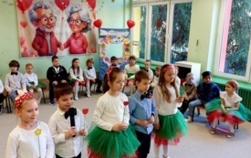 dzieci w odświętnych ubraniach siedzą na krzesełkach w p&oacute;łkolu za nimi dekoracja na dzień babci i dziadka piątka dzieci stoi na środku jedno z dzieci trzyma mikrofon