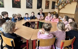 dzieci siedzą przy stolikach ustawionych w koło wśr&oacute;d nich siedzi pani trzymająca książkę