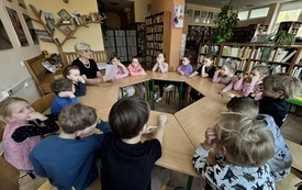 dzieci siedzą przy stolikach ustawionych w koło wśr&oacute;d nich siedzi pani trzymająca książkę
