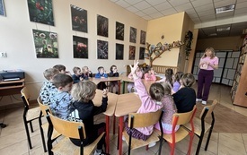dzieci siedzą przy stolikach ustawionych w koło przed nimi stoi panie trzymająca książkę