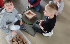 dzieci siedzą przy pudełku z drewnianymi klockami przed nimi budowla wykonana z tych klock&oacute;w ustawiona w okręgu