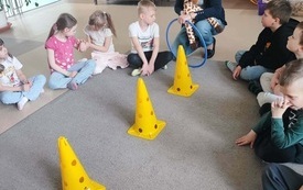 dzieci siedzą na dywanie na środku ustawione trzy ż&oacute;łte pachołki przed dziećmi kuca pani kt&oacute;ra trzyma pluszową żyrafę